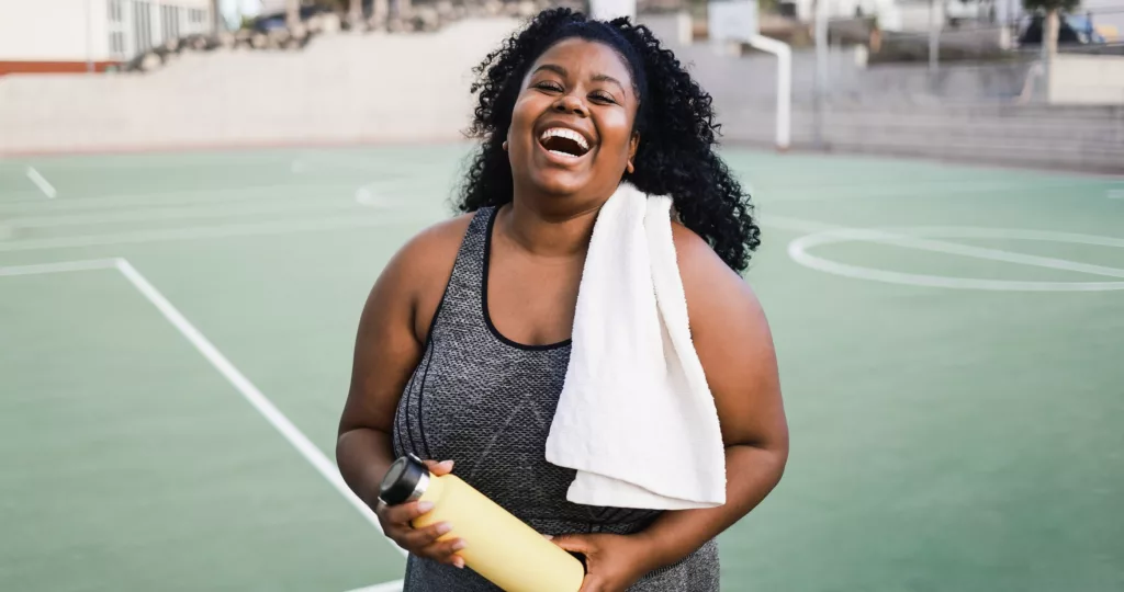Mulher rindo com toalha e garrafa de bebida após treino em uma quadra esportiva