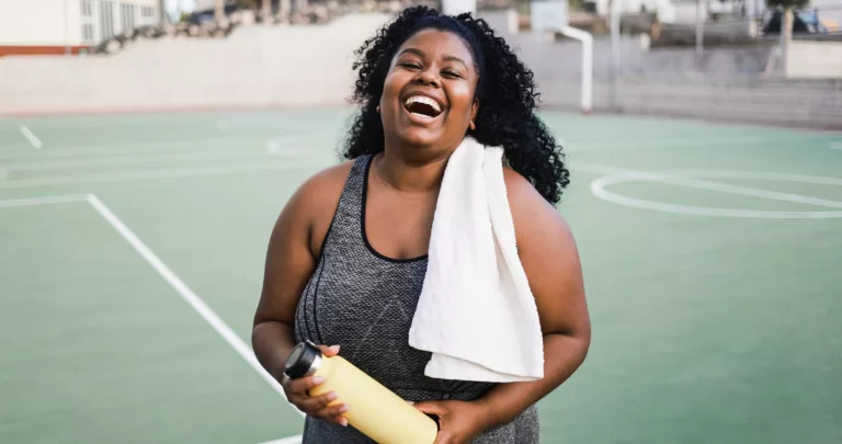 Mulher rindo com toalha e garrafa de bebida após treino em uma quadra esportiva