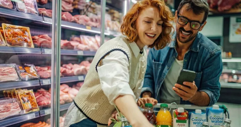 Casal consultando o celular diante da gôndola de carnes de um supermercado