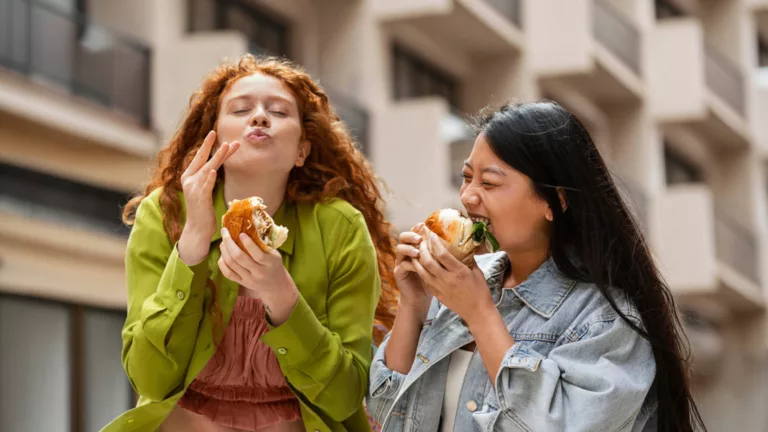 Duas jovens comendo sanduíches na rua, momento de consumo típico do foodservice